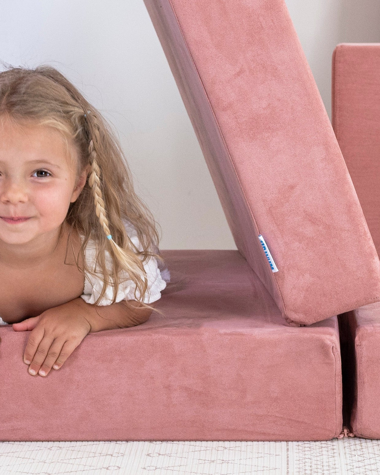 A young girl with blonde hair smiles while lying inside a pink foam play structure shaped like a triangle, next to a matching pink foam cushion and sofa on a white floor against a white wall.