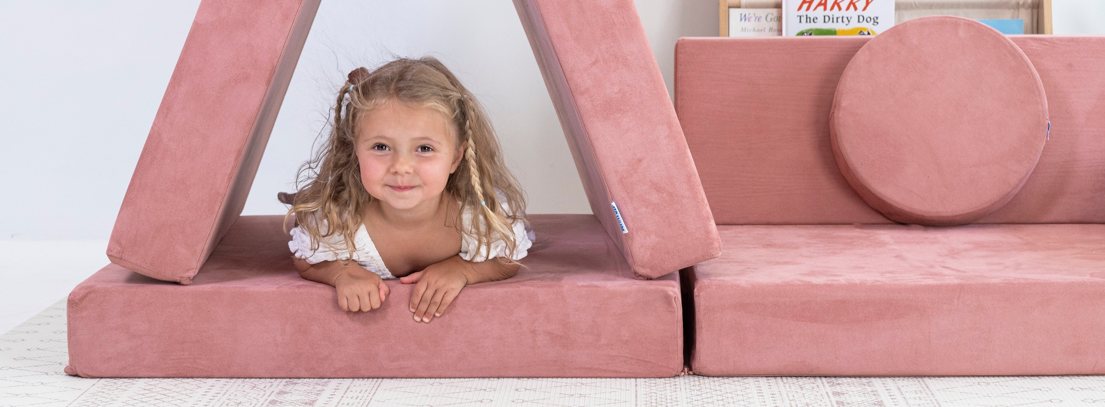 A young girl with blonde hair smiles while lying inside a pink foam play structure shaped like a triangle, next to a matching pink foam cushion and sofa on a white floor against a white wall.